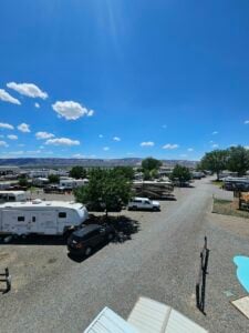 Wide view of a sunny RV park with several parked trailers and cars. The gravel pathways meander between green trees and neatly positioned vehicles. The horizon reveals a distant, rocky mountainous landscape under a bright blue sky with scattered clouds.