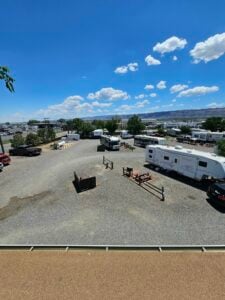 An expansive gravel lot on a sunny day with several RVs, trailers, and trucks parked in various spots. Some trees and utility poles are visible in the background, and the area is surrounded by a fence. Fluffy clouds dot the blue sky above.
