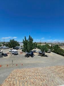 A rooftop view of an outdoor area with recreational vehicles (RVs) and cars parked on a gravel lot. Several trees provide greenery among the vehicles. Orange traffic cones are scattered in the foreground. In the distance, power lines run along the horizon under a clear blue sky.
