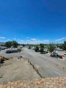 A campsite with scattered RVs and vehicles under a clear blue sky. The ground is gravelly with patches of dirt. There are trees and bushes around the site, and a few cones are visible in the distance. A small playground area is seen on the left.