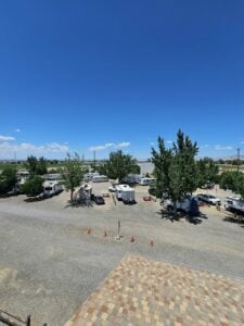 A clear, bright day at an RV park with several RVs and vehicles parked on gravel lots. Trees offer shade throughout the area, and there are orange traffic cones near the center. In the background, a vast landscape extends under a clear blue sky.