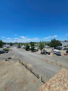 A gravel parking lot filled with several white RVs and trailers, surrounded by a few trees and picnic tables. The background features a green field, power lines, and a clear blue sky. Orange traffic cones are placed near the right side of the image.