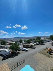 A scenic view of a large RV park on a sunny day. Various RVs and motorhomes are parked across the lot, with trees providing shade. The sky is clear with a few fluffy clouds, and mountains can be seen in the distant background. A playground is visible on the right side.