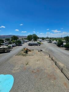 A gravel RV park under a clear blue sky, with various RVs parked in rows. There is a small playground area with a swing set and slide. Trees are scattered throughout the park, and mountains can be seen in the distance.