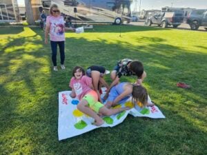 Four children are playing a game of Twister on a grassy lawn, each positioned on different colored dots, tangled up in playful poses. An adult woman stands nearby, holding the game's spinner, smiling and watching the kids. In the background, RVs and parked cars create a lively gallery of outdoor fun.