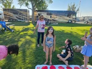 A young girl with face paint stands on the grass, surrounded by children. Adults and children are seen in the background near parked RVs and trees. A colorful Twister mat is on the ground. The scene appears to be a fun, gallery-like outdoor gathering with games.