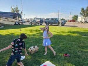 Two children are playing on a grassy area near parked vehicles and RVs. One child, wearing a blue and white dress, strikes a pose with one hand on her hip and the other holding a mask to her face. The other child, in a black shirt, is bending down near a cardboard box, creating an impromptu gallery of imagination.
