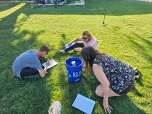 Three people are sitting on the grass, focused on papers. A blue bucket with "Do It Right" text is in the center. One person is writing, another is examining a sheet, and the third is reading. Nearby, a bundle of colorful straws adds a playful touch, like an outdoor art exhibition capturing creativity in action.