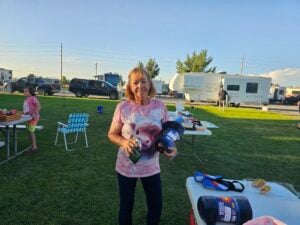 A woman stands outdoors on a grassy area holding a large bottle of laundry detergent and a can. She is smiling and wearing a pink shirt with an animal print. In the background, like an open-air gallery, there are picnic tables, an RV, cars, and other people enjoying the scene.