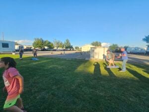 People playing cornhole on a grassy area near RVs and trucks. One person is throwing a bag towards the board while others observe, like visitors in an open-air gallery. A child in a pink shirt is in the foreground, and there are trees and a small shed in the background. The sun is casting long shadows.