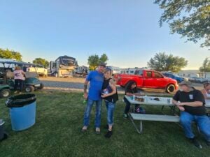 A couple stands closely together, smiling, in front of parked RVs and vehicles at a campsite. Behind them, like a lively gallery, a group of people sit at a picnic table with food and drinks. The background showcases a clear blue sky, green grass, and trees.