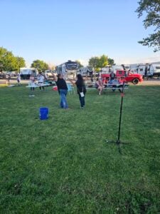 People are enjoying a sunny day at an RV park. Two individuals face each other, possibly playing a game or engaging in an activity. Several RVs are parked in the background, and other people are seated or standing around the green field, creating an informal art gallery atmosphere. A microphone stand is in the foreground.