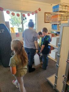 A group of children and an adult are standing in a room that doubles as a Halloween-themed art gallery. They are gathered around a display near shelves of boxes, examining the spooky exhibition. Sunlight is streaming in through a nearby window.