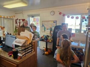 A bustling office buzzes with energy as two adults manage a counter brimming with papers, a computer, and a drink. Children weave through the festive Halloween decor, including banners and pumpkins. The scene resembles a lively gallery of autumnal celebration and activity.