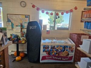 A small gallery corner of the store is decorated for Halloween. A coffin-shaped object labeled "RIP" stands next to an ice cream freezer. Holiday decorations include a banner with smiling pumpkins and two pumpkins by a table adorned with toys and maps.