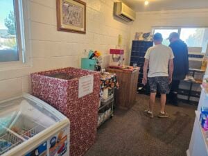 A small gallery-like room with two people standing near a corner. It features a popcorn machine, a freezer stocked with ice cream, and a large bin draped in patterned cloth. Shelves hold various items, including a framed picture and a potted plant adding charm to the space.