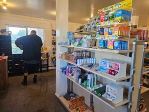 A quaint store interior resembles a charming gallery, with shelves showcasing personal care items, snacks, and over-the-counter medications. A person stands near a window in the background. The room is brightened by a blend of natural and artificial light.