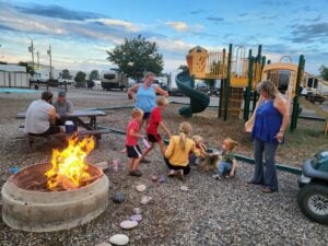 People enjoying a campground with picnic tables and a fire pit, creating a picturesque outdoor gallery. Children play near a playground while adults oversee. The sky is a mix of blue and clouds, and RVs are parked in the background.