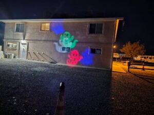 A building at night becomes a vibrant gallery, with colorful ghost illustrations projected onto its walls. In the foreground, a blurred orange cone adds depth, while a tree and parked vehicles create a tranquil backdrop.