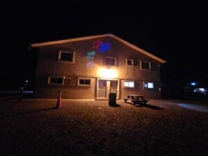 A two-story gallery is lit by a central porch light at night, casting colorful patterns on its upper facade. In the foreground on a gravel surface, a wooden picnic table and an orange traffic cone add contrasting textures and colors to the scene.