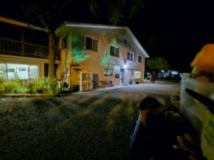 A two-story building at night with exterior lights illuminating its walls, resembling an art gallery. The scene is dimly lit, with a tree on the left and parked vehicles on a gravel driveway. The upper floor features multiple windows, showcasing glimpses of creativity within, and there's some greenery nearby.
