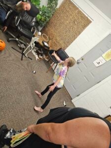 A girl in a tie-dye shirt and black pants is playing indoors, tossing rings toward a skeleton decoration hanging near the gallery wall. Two adults are in the room, one holding additional rings and another seated. A pumpkin and a plant add to the festive atmosphere in the background.