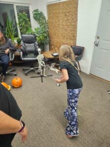 A young girl with long hair wearing a black shirt and floral pants prepares to throw a ring in a room resembling an art gallery, adorned with Halloween decorations including carved pumpkins and a skeleton. Two people are seated nearby, surrounded by vibrant plants and a bamboo curtain.