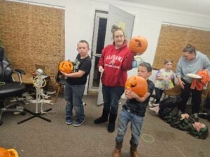 In a cozy indoor setting, a vibrant gallery of carved pumpkins is proudly displayed by both children and adults. Amidst the festive scene, one person sports a red Alabama hoodie, while others showcase pumpkins with unique designs. A skeleton decoration adds to the spooky atmosphere in the background.