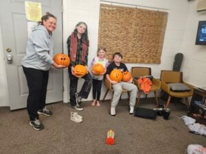 In a cozy indoor setting, resembling an art gallery of creativity, three children and an adult proudly display their carved pumpkins. Chairs flank a TV, with a door displaying a notice nearby. A skateboard casually leans against the wall as the barefoot kids enjoy this festive moment.