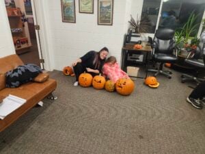 A woman and a child sit on the floor next to a gallery-like line of carved pumpkins. The woman smiles warmly, both dressed casually. In the background, a brown sofa and chairs are adorned with decorative items, creating a cozy atmosphere.