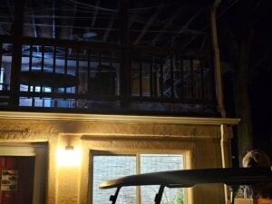 A dimly lit outdoor scene at night reveals a building with a balcony, reminiscent of an eerie gallery. The balcony, adorned with spider webs and bathed in soft yellow light, features a round table. Majestic trees frame the background, enhancing the mysterious allure.