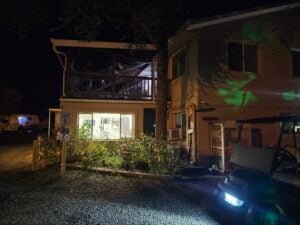 A dimly lit building at night, with a porch on the upper level and lights glowing inside like an inviting gallery. A golf cart is parked in front, its headlights cutting through the darkness. Small plants and a parking sign adorn the entrance, while gravel crunches underfoot.