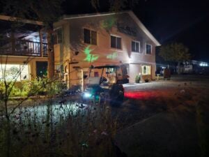 A golf cart with headlights on is parked in front of a two-story building at night. The building, resembling an art gallery, is illuminated with various light patterns, and there are trees and a lit display window nearby.