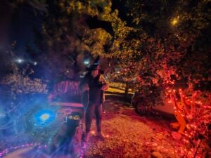 A person wearing a witch hat stands in a Halloween-themed yard at night, creating an eerie gallery of colorful lights and decorations. A cauldron bubbles amid trees with orange and purple lights, while a foggy effect drifts through the captivating scene.