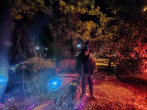 A person dressed as a witch stands in a spooky garden at night, resembling a gallery of Halloween frights. The scene includes a cauldron, spider web, bat decoration, and colorful lights, creating an eerie ambiance with glowing orbs and shadows.