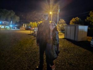 Two people stand together outside at night near a shed, illuminated by bright lights that cause lens flare. One person appears to be wearing a Halloween mask.