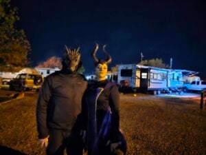 Two people in costumes, one wearing a dragon mask and black outfit, the other dressed as Maleficent, stand outdoors at night near trailers and parked vehicles.