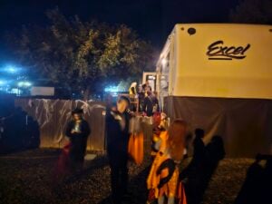 People dressed in costumes gather around a gallery-like RV adorned with shiny cobwebs at night, possibly for Halloween. The scene is lit with a warm, ambient glow as individuals clutch bags or buckets.