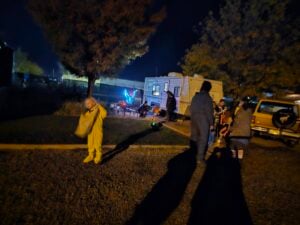 Children and adults in costumes stand near a parked trailer at night, with trees and a lit outdoor area visible in the background.