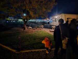 A toddler in an orange jacket walks near adults at a dark campsite, resembling a scene from a gallery. A tree with autumn leaves stands nearby, and a lit white RV glows in the background. The setting is dimly illuminated, suggesting evening or nighttime.