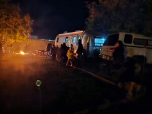 A nighttime scene unfolds like a gallery of nature with a group gathered around a flickering campfire. An illuminated camper and parked truck add charm, while trees and picnic tables form the perfect backdrop.