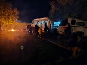 In a nighttime outdoor scene, people gather around a campfire near a silver trailer and a white truck, creating an impromptu exhibition of warmth. Trees and picnic tables frame this living gallery, with the fire's glow illuminating the area like artwork on display.