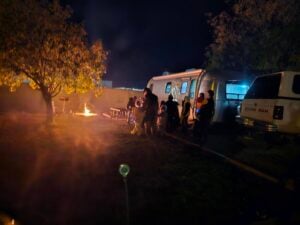 A group of people gathers like a cozy gallery around a campfire near a parked caravan and truck at night. Chairs and a tree stand nearby, with warm, glowing lights illuminating the scene under the dark sky.