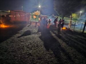 A group of people gathered outside at night, illuminated by nearby fires like a lively outdoor gallery. Shadows stretch across the gravel ground, while in the background, trees and structures are dimly visible under streetlights.