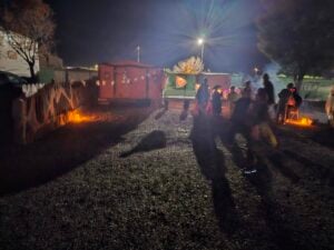 A nighttime scene with silhouettes of people gathered in a gravel area, delicately illuminated by gallery-style soft lights. Halloween decorations adorn nearby structures, casting playful shadows on the ground and creating a festive atmosphere.