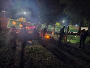 A dimly lit pathway at night features people strolling under the glow of streetlights. Halloween pumpkin decorations hang on a fence, casting an eerie atmosphere like an open-air gallery of shadows formed by trees and passersby.