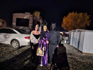 Three people in costumes stand outdoors at night, forming a captivating gallery. The first person is dressed in black with a shawl, the second sports a purple and black outfit, and the third wears a dark ensemble with a dragon mask. A car and camper are silhouetted in the background.