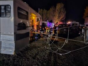 A group of people gather outside a lit RV at night, with ropes strung across the area and vehicles parked nearby under illuminated trees.