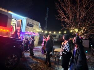 People stand outside a camper decorated with colorful lights at night; some talk while others enter the camper, and string lights are hung in the background.