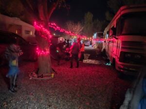 A row of RVs and a tree decorated with red string lights at night, with people gathered and walking on a gravel area.